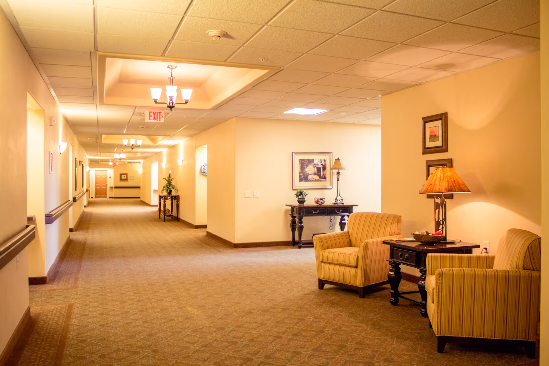A well-lit hallway in a senior living facility with beige walls and carpeted floors. On the right side, there are two striped armchairs with a small wooden table and a lamp between them. Further down the hallway, there are decorative tables with plants and framed pictures on the walls. Ceiling lights and an exit sign are visible.