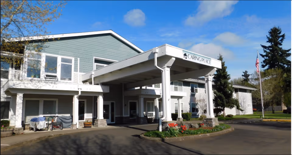 Front entrance of a two-story senior living facility with a covered drop-off canopy, landscaped planters, and an American flag.