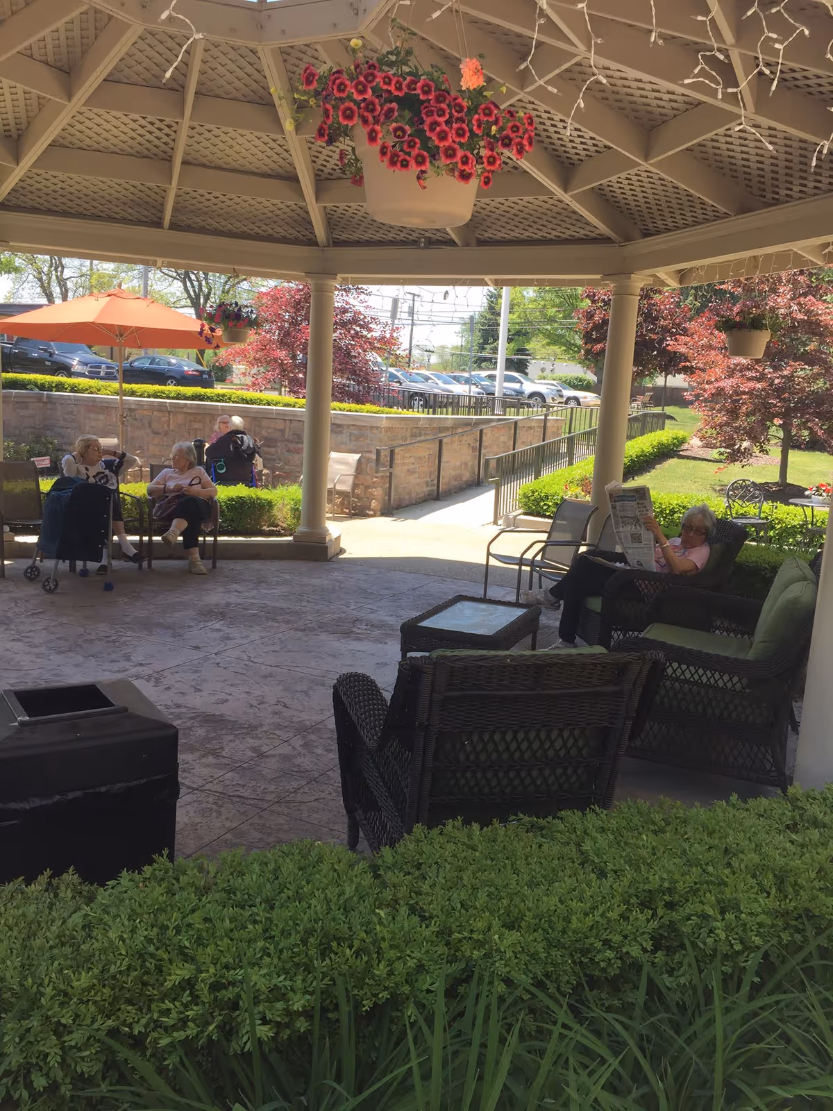 Outdoor covered patio area with several senior residents sitting on chairs and benches. There are hanging flower pots with pink flowers, an orange umbrella, and greenery surrounding the patio. A woman is reading a newspaper while others are engaged in conversation.