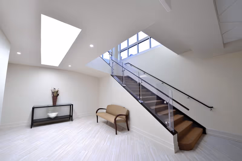 Bright interior space with a staircase featuring glass and metal railings, a beige cushioned bench with wooden armrests, a black console table with a decorative vase and bowl, and a large skylight overhead.
