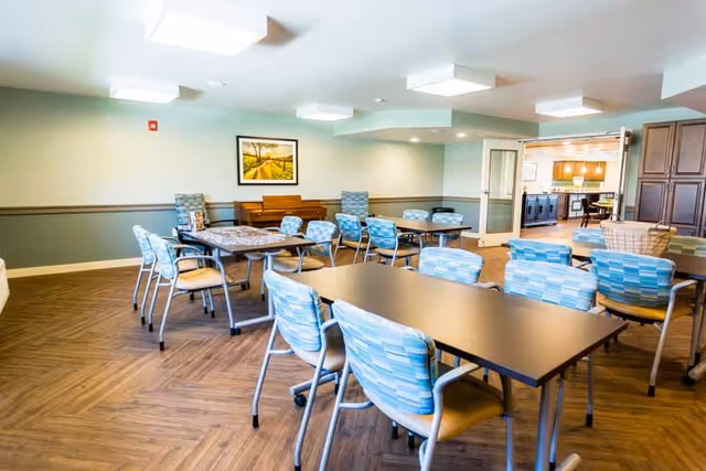 A bright and spacious common area with multiple tables and blue patterned chairs arranged neatly. The room has wooden flooring, soft green walls with a chair rail, and a framed picture above a piano against the far wall. In the background, there is an open doorway leading to a kitchen area with dark cabinetry and pendant lighting.