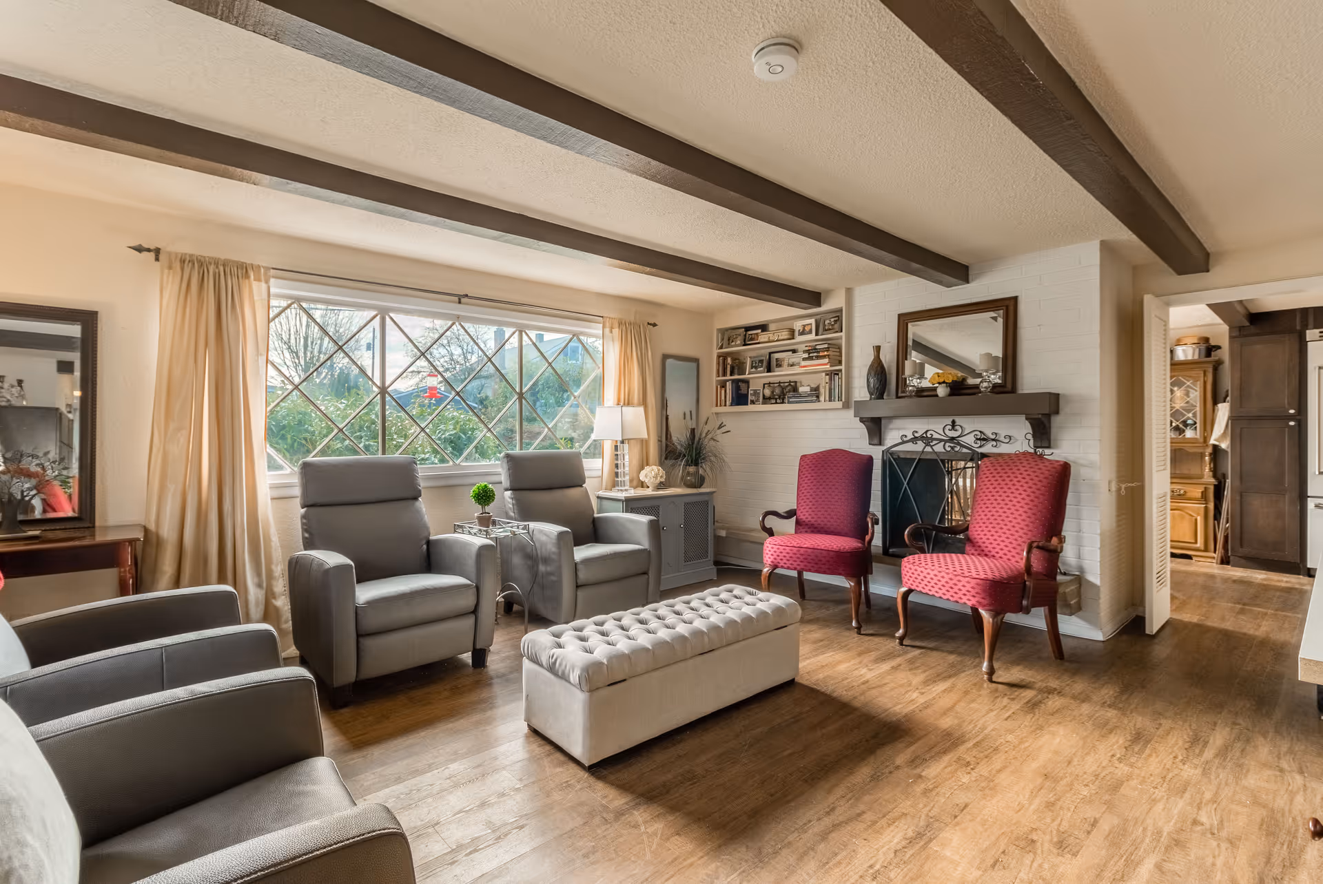 Sunlit living room with gray recliners, two red armchairs by a fireplace, a tufted ottoman, exposed ceiling beams and a large diamond-pane window.