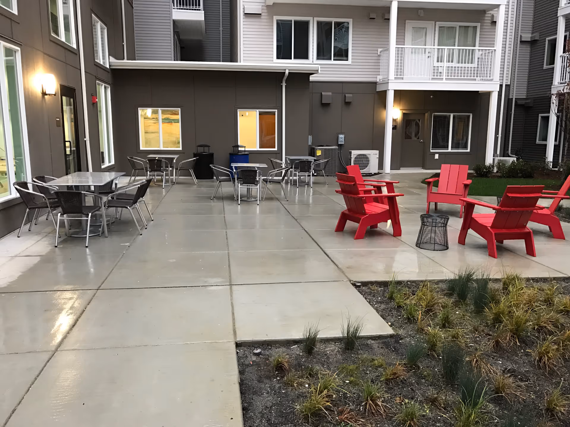 Outdoor patio area at the Reserve at SeaTac featuring several metal tables with chairs on a concrete surface, and a group of red Adirondack chairs arranged around a small black metal side table. The patio is adjacent to a multi-story building with windows and balconies, and there is some landscaping with grass and plants in the foreground.