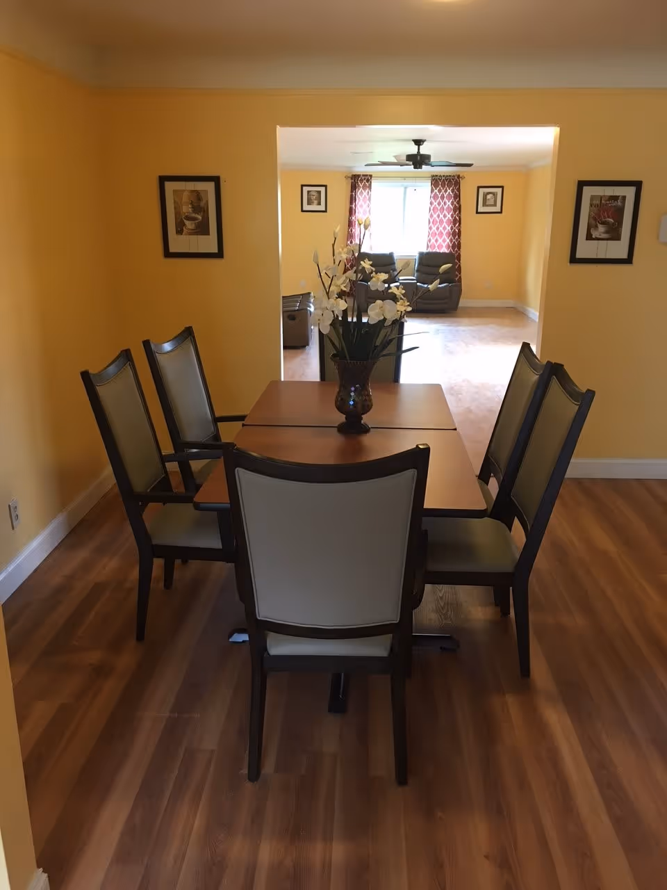 A dining area with a wooden table and six chairs arranged around it. A vase with white flowers is placed in the center of the table. The walls are painted yellow, and there are framed pictures hanging on the walls. In the background, there is a living room area with two recliner chairs, a window with red patterned curtains, and a ceiling fan.