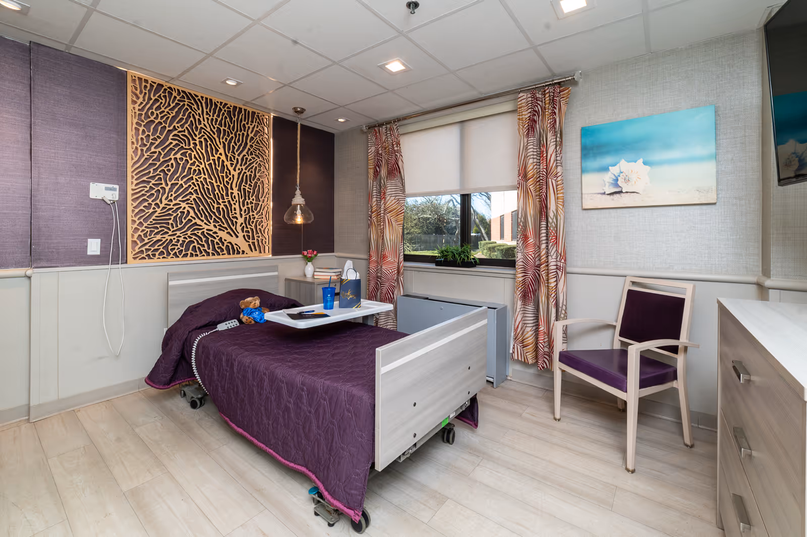 A well-lit nursing facility bedroom with a single hospital-style bed covered in a purple quilt. A tray table on the bed holds a blue cup, a remote control, and a small teddy bear. The room features a decorative wooden wall panel, patterned curtains, a window with a view of greenery outside, a purple cushioned chair, a dresser, and a wall-mounted TV.