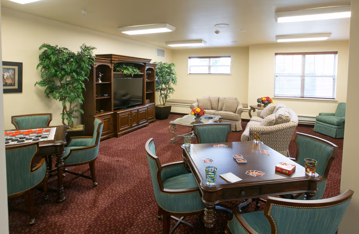 A cozy common living area in a retirement facility featuring a large wooden entertainment center with a flat-screen TV, two patterned sofas, a green armchair, and two tables with green upholstered chairs. One table has a checkers game set up, and the other has playing cards and glasses. The room has beige walls, red patterned carpet, two windows with blinds, and decorative plants.