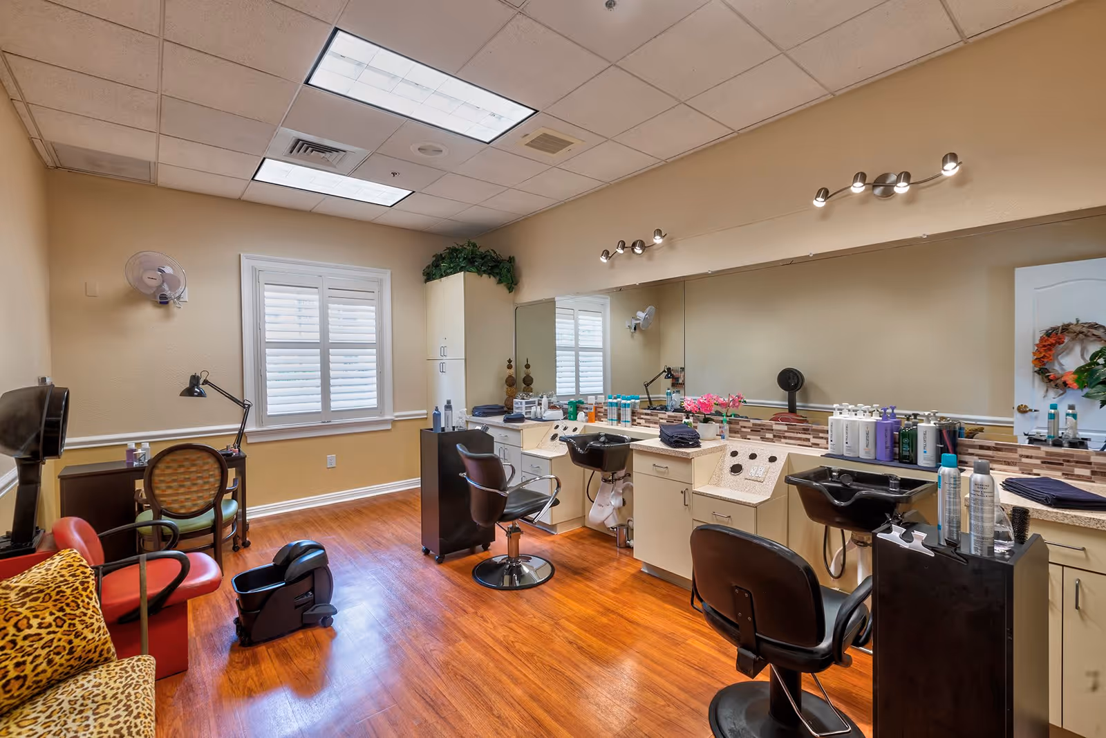 Interior view of a salon room in a senior living facility with wooden flooring, salon chairs, hair washing stations, a large mirror, and various hair care products on the counter. There is a window with white shutters, a small desk with a chair, and a leopard print cushioned chair in the corner.