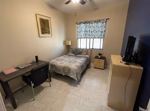 A small furnished bedroom with a bed, nightstand, dresser with TV, desk and chair, patterned curtains and tiled floor.