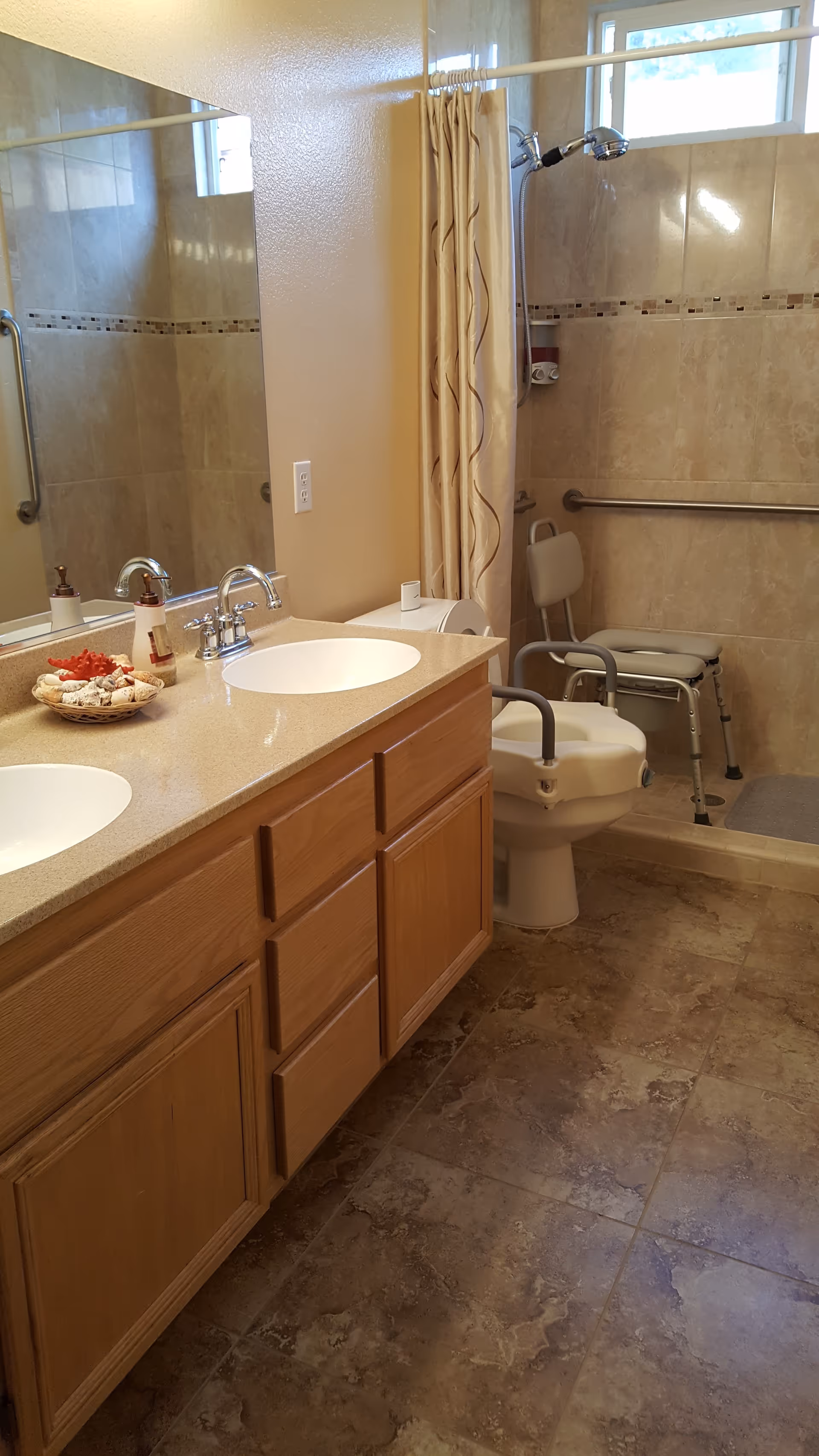 A bathroom with a double sink vanity featuring wooden cabinets and a beige countertop. There is a large mirror above the sinks. The bathroom has a tiled floor and walls with a decorative tile border. A shower area with a curtain, grab bars, and a shower chair is visible. There is also a toilet with an attached raised seat and armrests for accessibility.