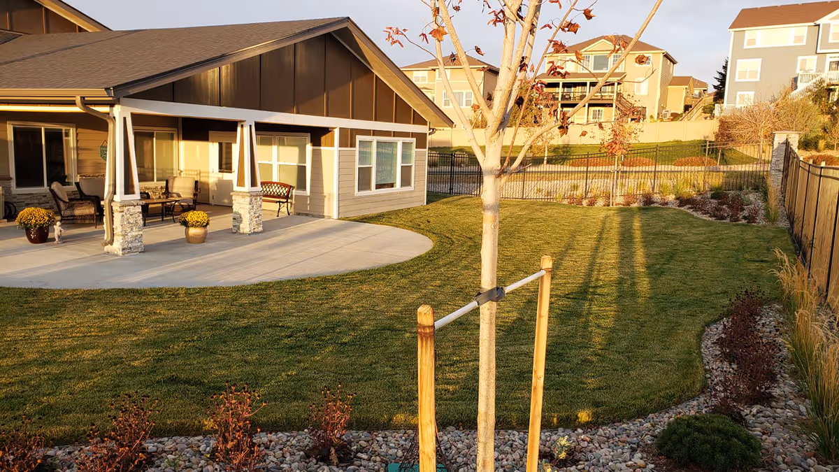 A well-maintained outdoor garden area of a residential facility with a small tree supported by wooden stakes in the foreground, a green lawn, landscaped flower beds with rocks, and a covered patio attached to a beige and brown building with outdoor seating. Other residential buildings are visible in the background under a clear sky.