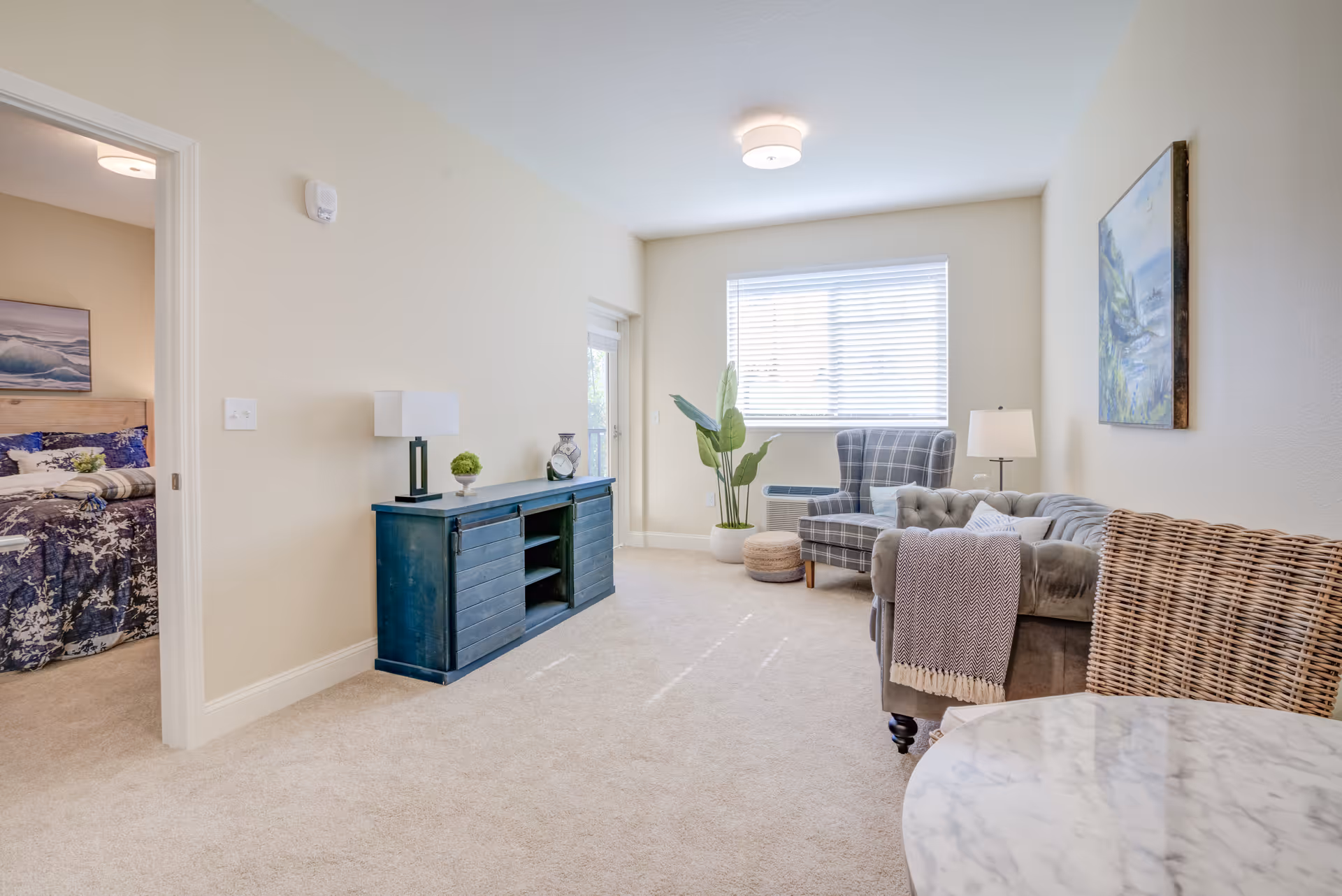 A bright living room with beige walls and carpet, featuring a gray tufted sofa with a throw blanket, a plaid armchair, a wicker chair, a blue wooden cabinet with decorative items, a potted plant, a floor lamp, and a window with blinds. A bedroom with a bed and ocean-themed artwork is visible through an open doorway.