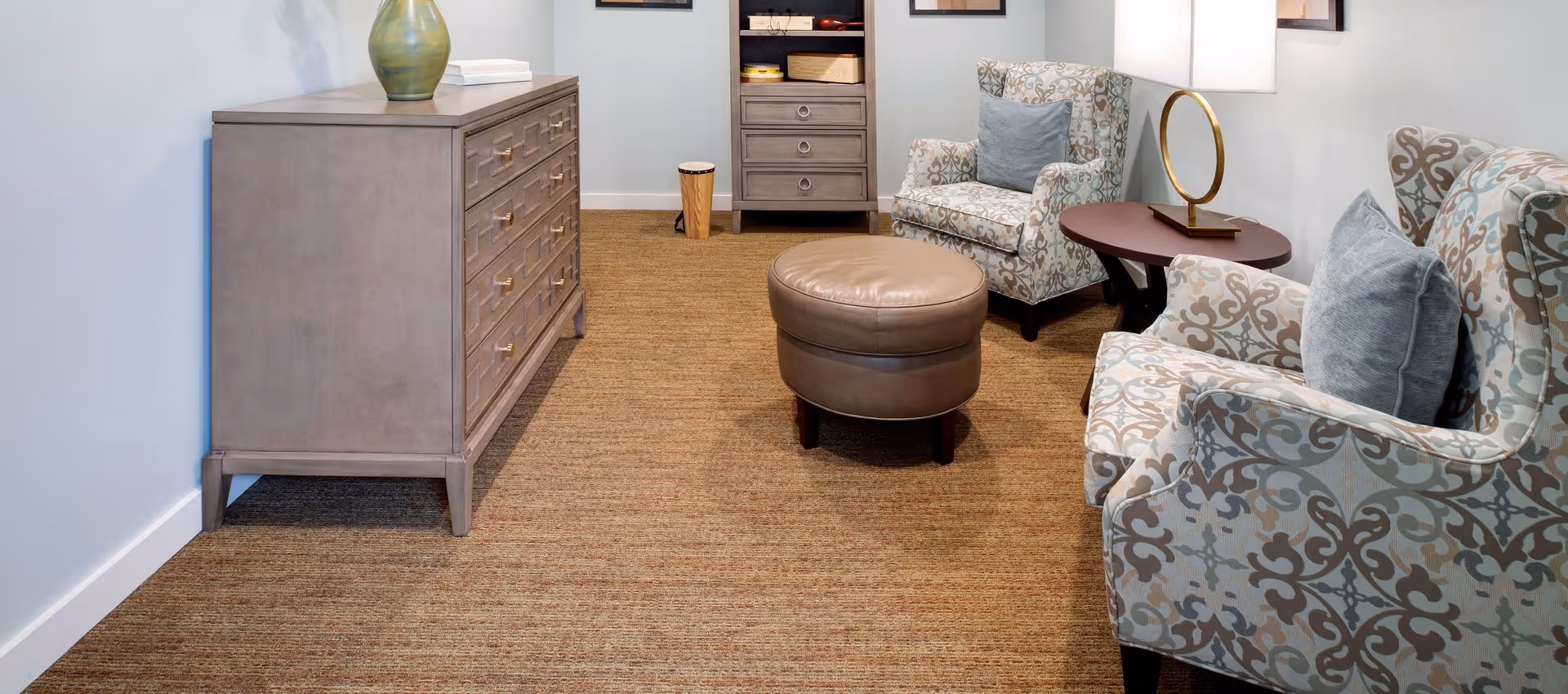 A cozy sitting area with two patterned armchairs with blue cushions, a round brown leather ottoman, a wooden side table with a lamp, a tall dresser with a green vase on top, and a smaller cabinet with drawers against a light-colored wall.