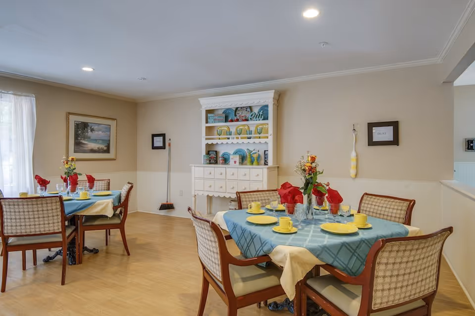 A dining room with two tables set for a meal. Each table has a blue tablecloth with a cream-colored underlay, yellow plates and cups, and red napkins. There are wooden chairs with cushioned seats around the tables. A white hutch with decorative plates and items is against the wall. The room has beige walls, a framed painting, and a broom hanging on the wall.