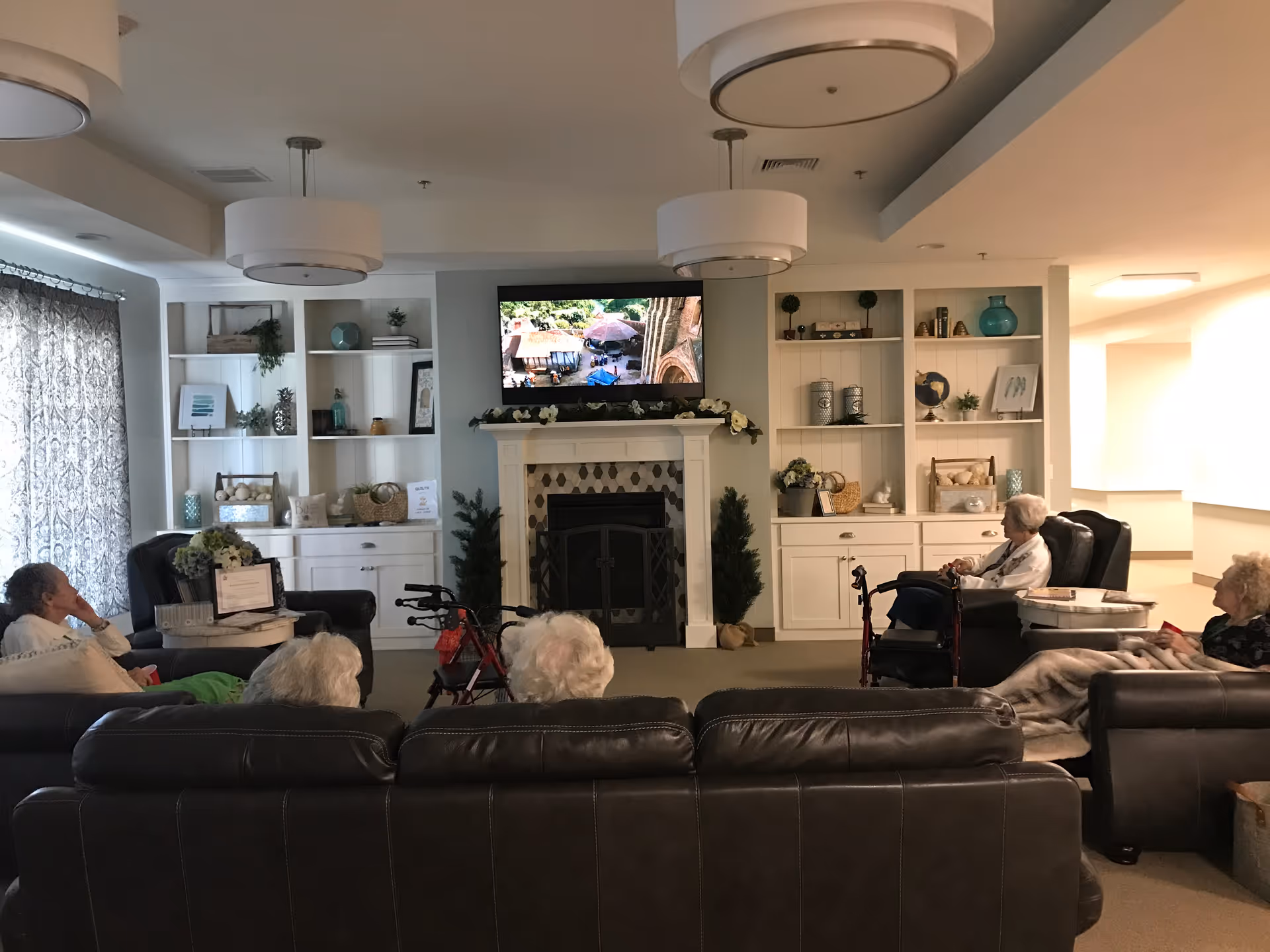 A cozy common living room with leather sofas and armchairs facing a TV above a fireplace, built-in white shelving, and several seated residents.