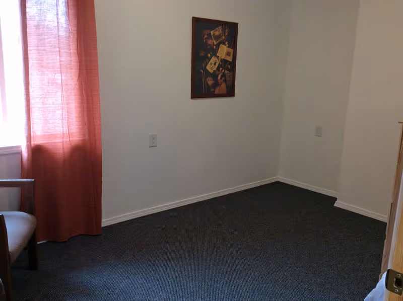 A small empty room with white walls and dark carpet. There is a window with a red curtain on the left side, a wooden chair partially visible, and a framed picture hanging on the wall. The room appears to be simple and minimally furnished.