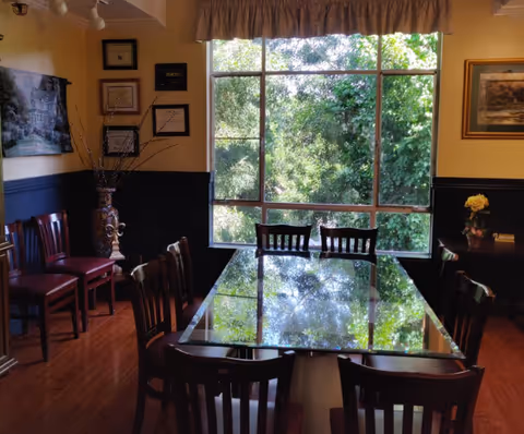 A dining room with a large glass-top table surrounded by wooden chairs. The room has a large window showing green trees outside, framed by a valance. The walls are decorated with framed pictures and a vase with branches is placed in the corner.