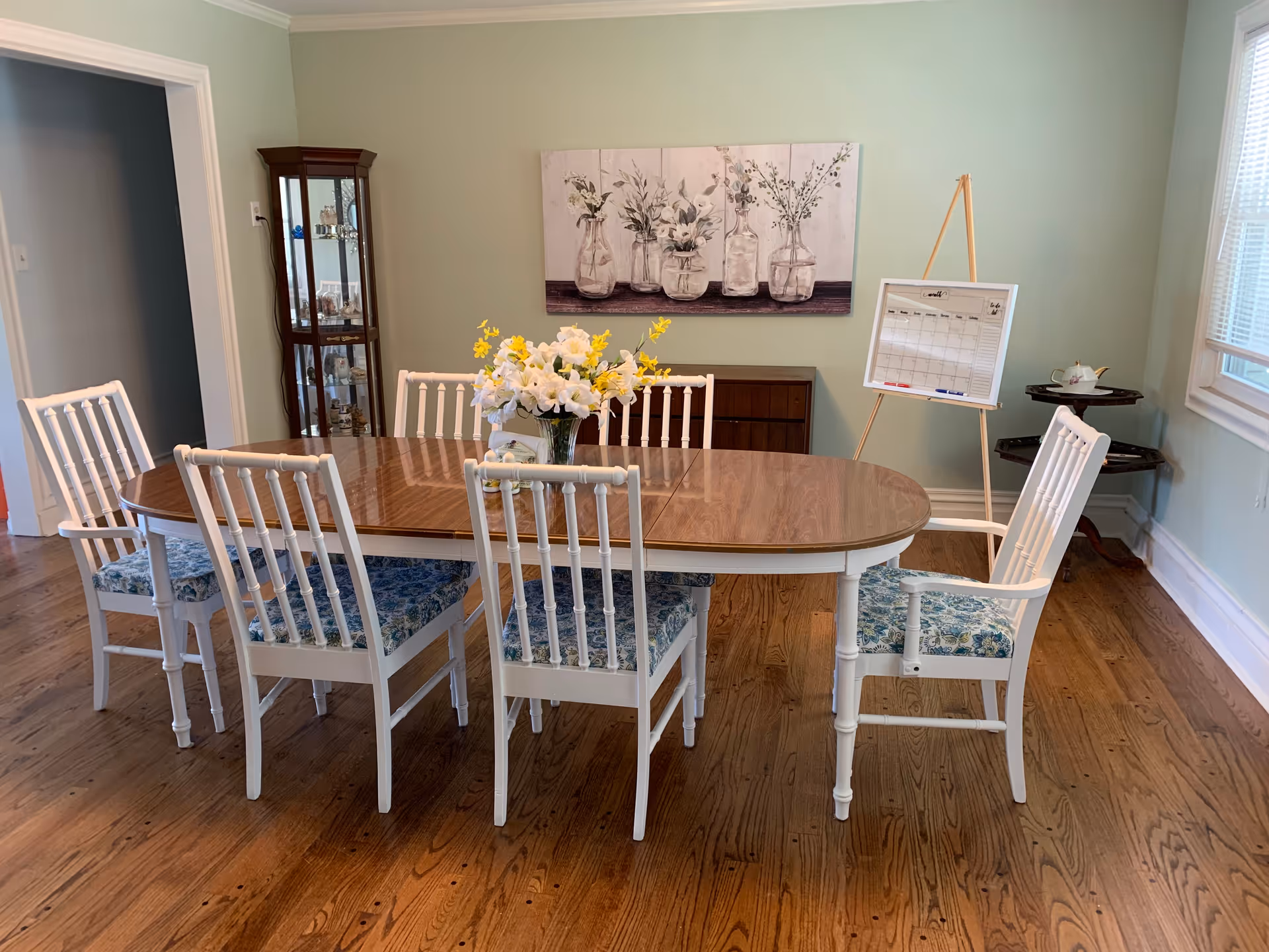 A dining room with a wooden table and six white chairs with blue patterned cushions. A vase with white and yellow flowers is placed on the table. On the wall behind the table, there is a painting of vases with flowers. To the right, there is a whiteboard on an easel and a small corner table with a teapot. The room has wooden flooring and light green walls.