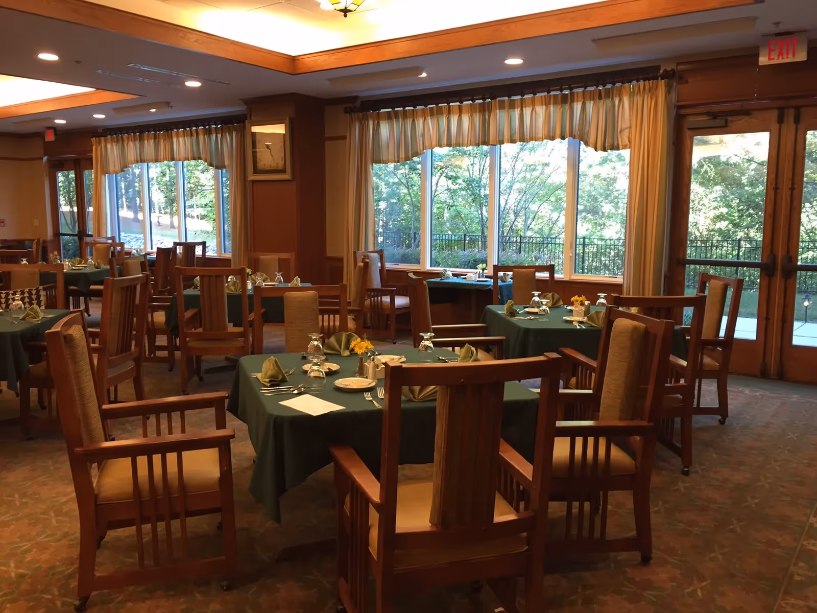 Dining room with multiple tables covered in green tablecloths, each set with plates, napkins, glasses, and silverware. Wooden chairs with cushioned seats surround the tables. Large windows with curtains allow natural light to brighten the room and offer a view of trees outside.