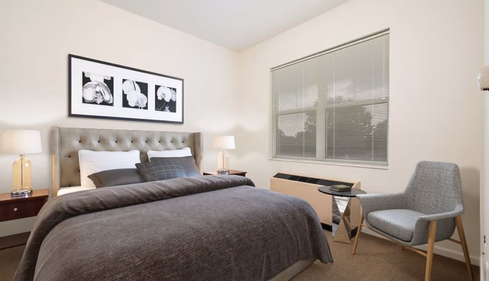 A cozy furnished bedroom with a tufted headboard, bedside lamps, a gray bedspread, artwork above the bed, and a window with blinds next to a chair and small table.