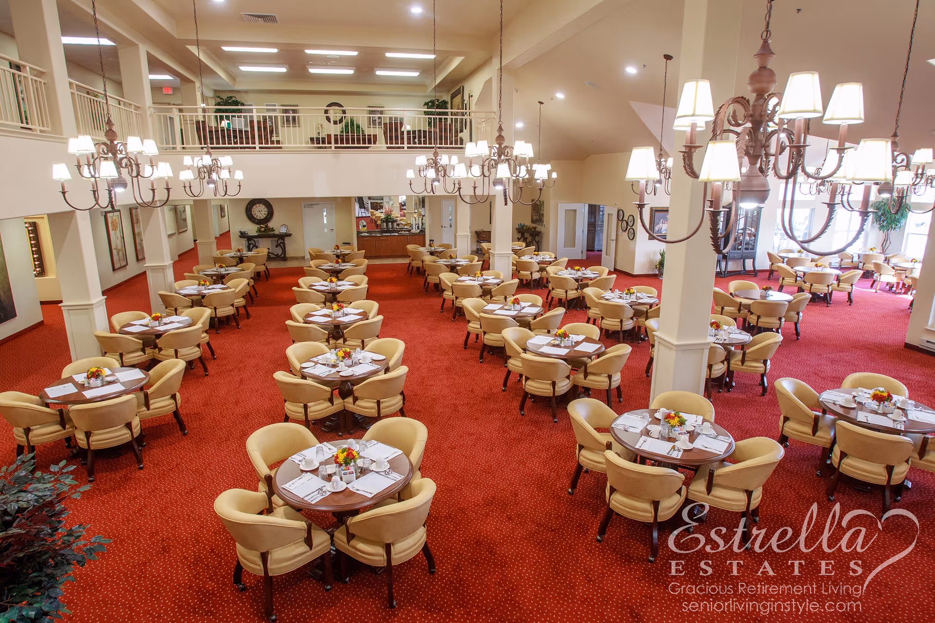 Spacious dining room with rows of round tables set for meals, beige chairs, red carpet, chandeliers, and a mezzanine.