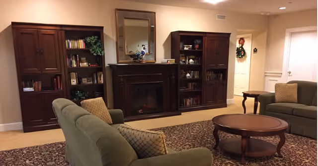 A cozy living room with two green upholstered sofas facing each other, a round wooden coffee table in the center, and a patterned area rug underneath. The back wall features a dark wooden fireplace mantel with a mirror above it, flanked by two tall wooden bookshelves filled with books and decorative items. The room has beige walls and carpet, with a small side table next to one sofa and a wreath hanging on a door in the background.