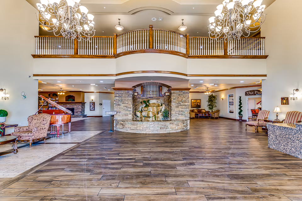 Spacious senior living facility lobby with wooden flooring, a stone water feature in the center, elegant chandeliers, a grand piano on the left, and comfortable seating areas with armchairs and small tables on both sides. The upper level features a wooden railing balcony overlooking the lobby.