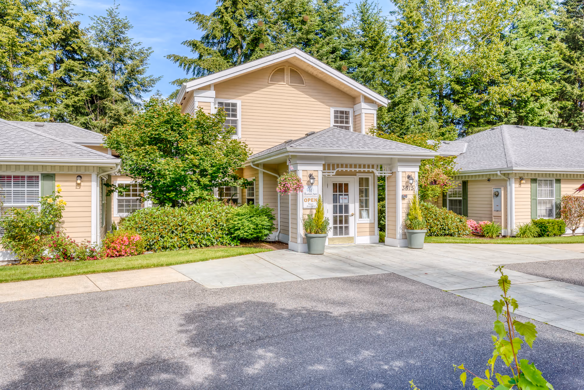 Exterior view of Hearthside Manor Memory Care building with beige siding, white trim, and a covered entrance. The building is surrounded by green trees, bushes, and colorful flowers under a clear blue sky.