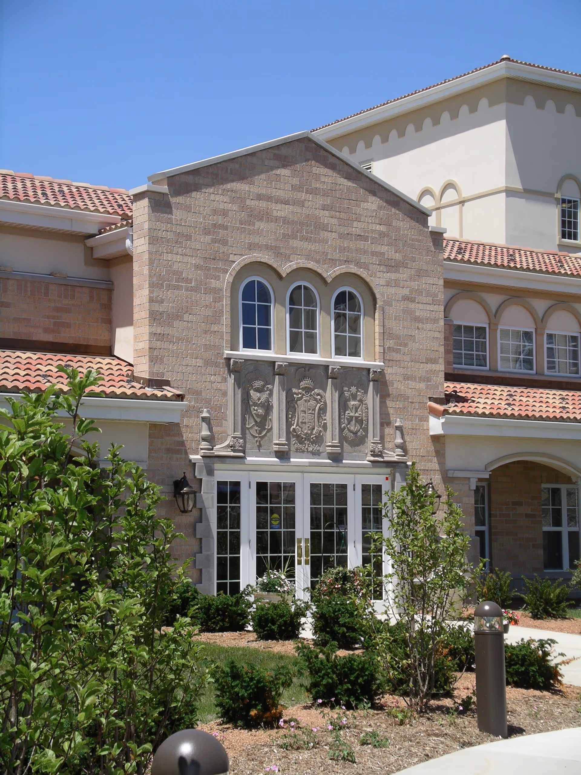 Exterior view of St. Catherine Commons - Independent Living building with beige brick facade, arched windows, decorative stone plaques above the entrance, and landscaped greenery in front under a clear blue sky.