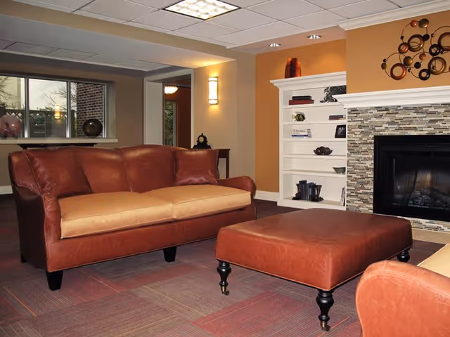 A cozy living room area featuring a brown leather loveseat and matching ottoman on a carpeted floor. In the background, there is a white built-in bookshelf with decorative items and a fireplace with a mosaic tile surround. The walls are painted in warm tones, and a modern wall sconce provides lighting.