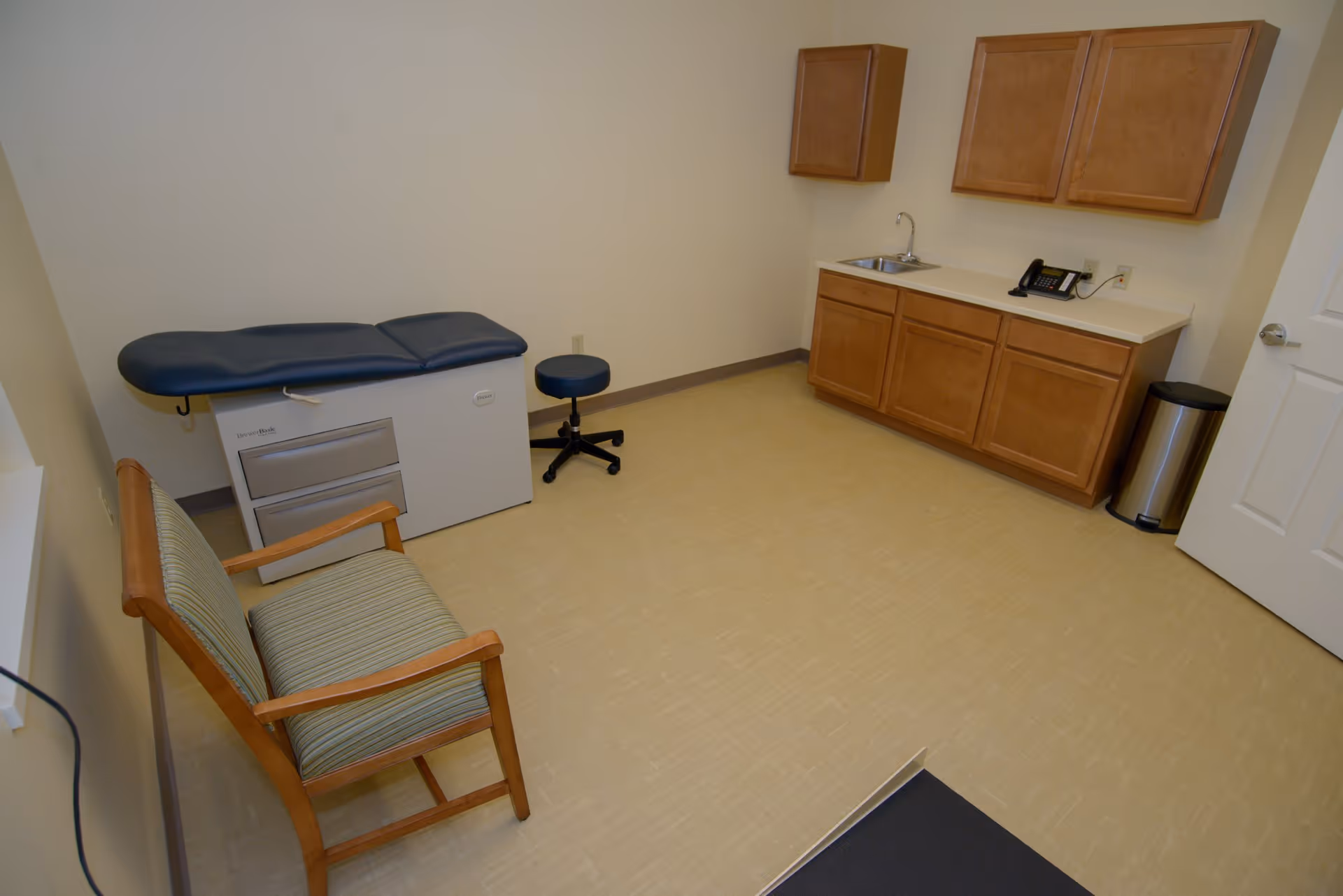A small medical examination room with a padded examination table, a rolling stool, a wooden chair with striped upholstery, wooden cabinets with a countertop and a sink, a telephone, and a trash can. The room has beige walls and flooring.