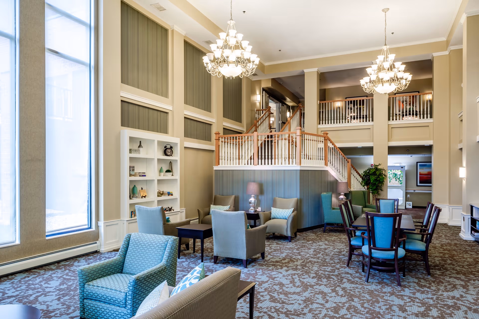 Spacious two-story senior living common area with seating, dining tables, chandeliers, and a staircase leading to an upper balcony.