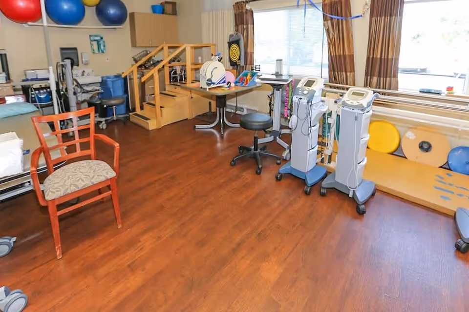 A rehabilitation therapy room with wooden flooring, exercise equipment including therapy balls, a small set of wooden stairs, medical machines on wheels, a wooden chair with a cushioned seat, and a window with brown curtains letting in natural light.