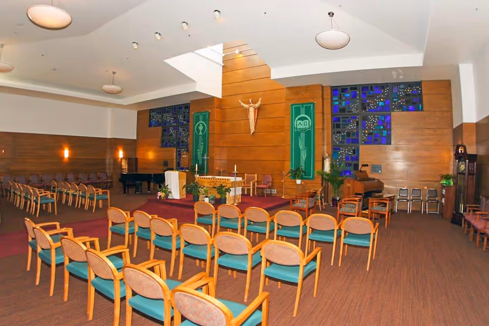 Interior view of a chapel or worship room with rows of wooden chairs with green cushions facing an altar. The altar is on a raised platform with religious decorations including a crucifix on a wooden-paneled wall, green banners, and stained glass windows. The room has a high ceiling with round hanging lights and additional seating along the walls.
