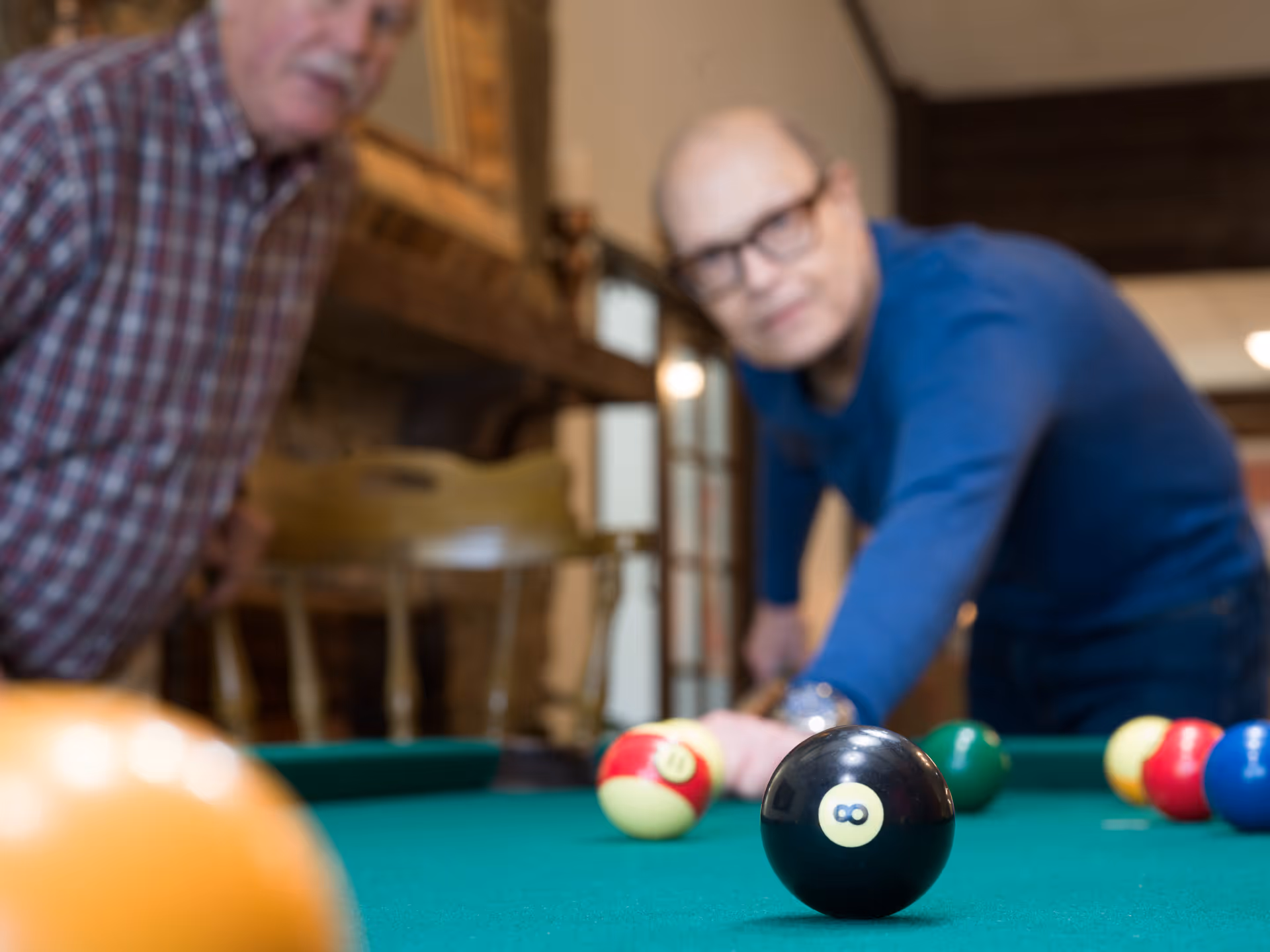 Two elderly men playing pool indoors, one man is aiming to hit the cue ball while the other watches. The focus is on the black 8-ball on the pool table.