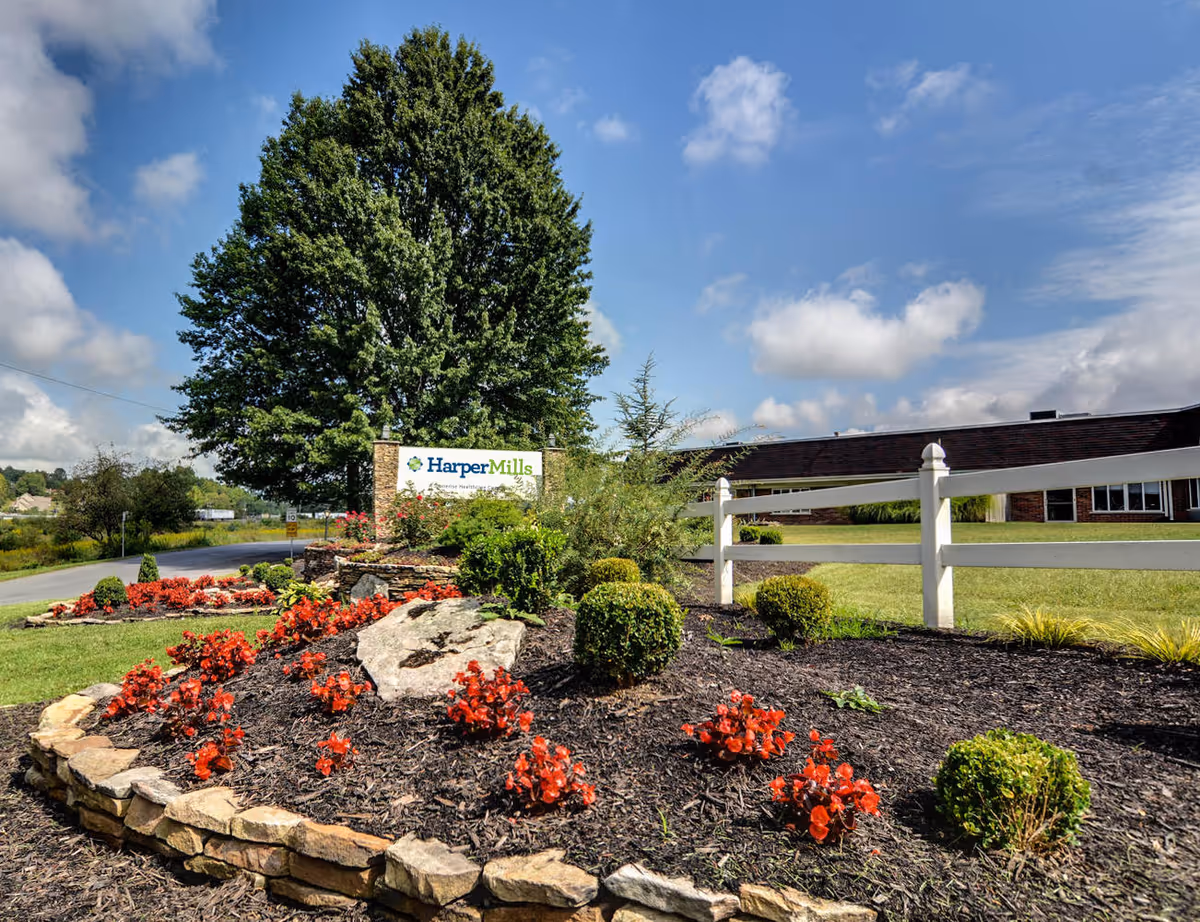 Outdoor landscaped garden area with red flowers, trimmed bushes, a large tree, and a white fence in front of a brick building under a partly cloudy blue sky. A sign reading 'Harper Mills' is visible near the tree.