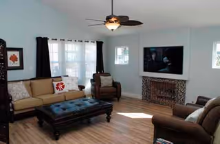 A cozy living room with a beige sofa, two brown armchairs, a large black ottoman coffee table, a ceiling fan with light, and a flat-screen TV mounted above a decorative fireplace. The room has light-colored walls, wood flooring, and windows with white curtains and dark drapes.