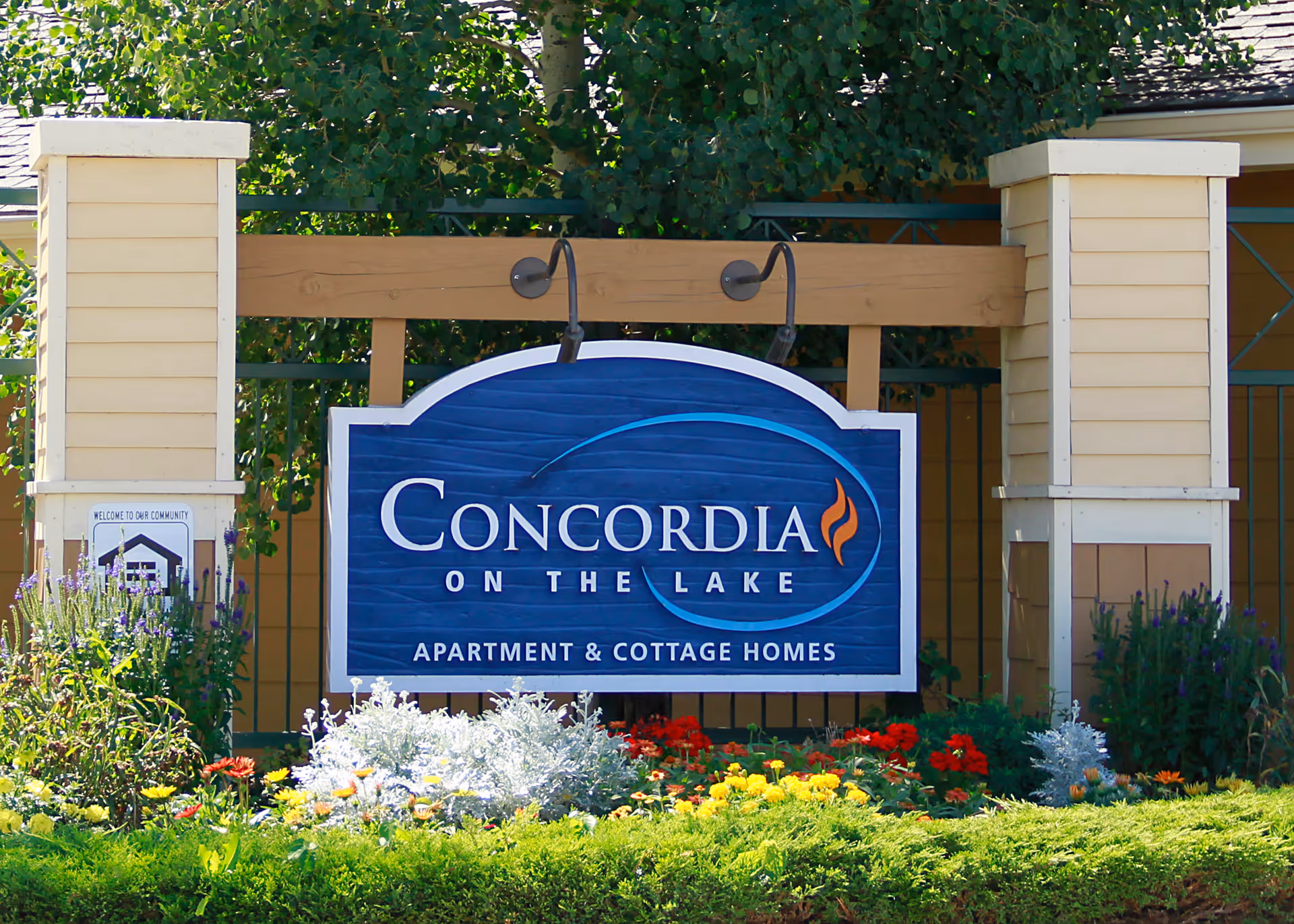 A large blue and white sign for Concordia On The Lake Apartment & Cottage Homes mounted on a wooden frame with two pillars on either side, surrounded by colorful flowers and greenery.