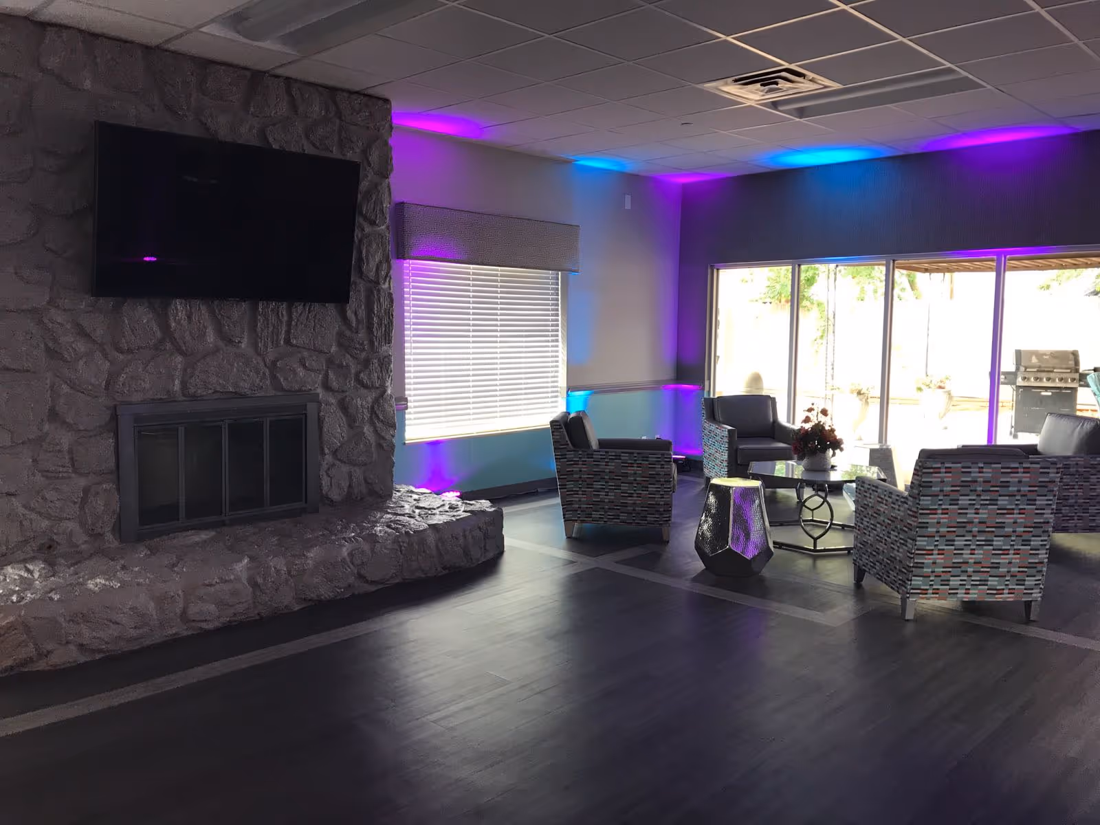A cozy living room area with a stone fireplace and a mounted flat-screen TV above it. The room features modern seating with patterned armchairs arranged around a glass coffee table. There is ambient purple and blue lighting along the walls and ceiling. Large windows and sliding glass doors let in natural light and provide a view of an outdoor patio with a grill.