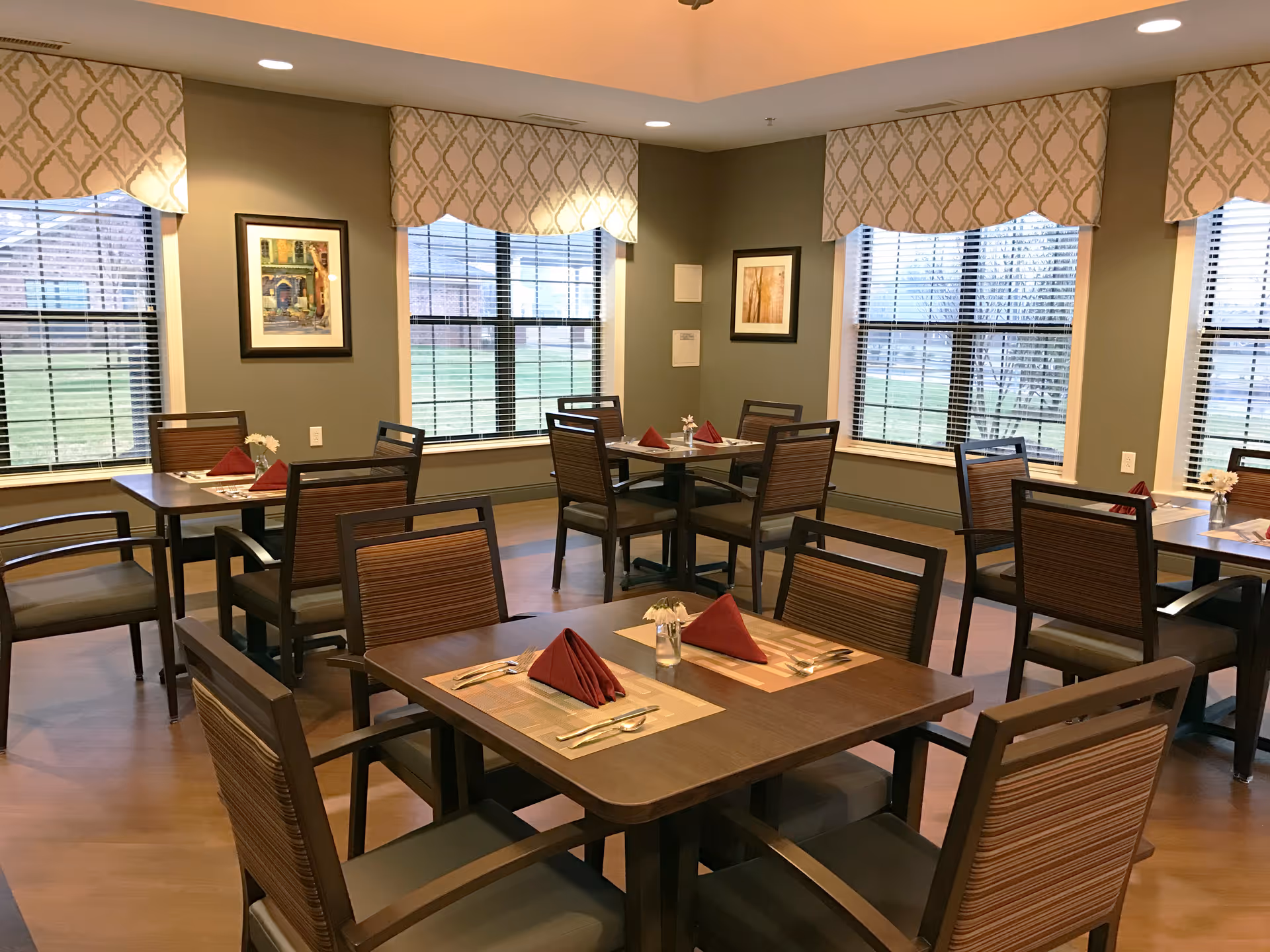 A dining room with several wooden tables and chairs arranged neatly. Each table has placemats, silverware, and folded red napkins. The room has large windows with patterned valances, framed artwork on the walls, and wooden flooring.