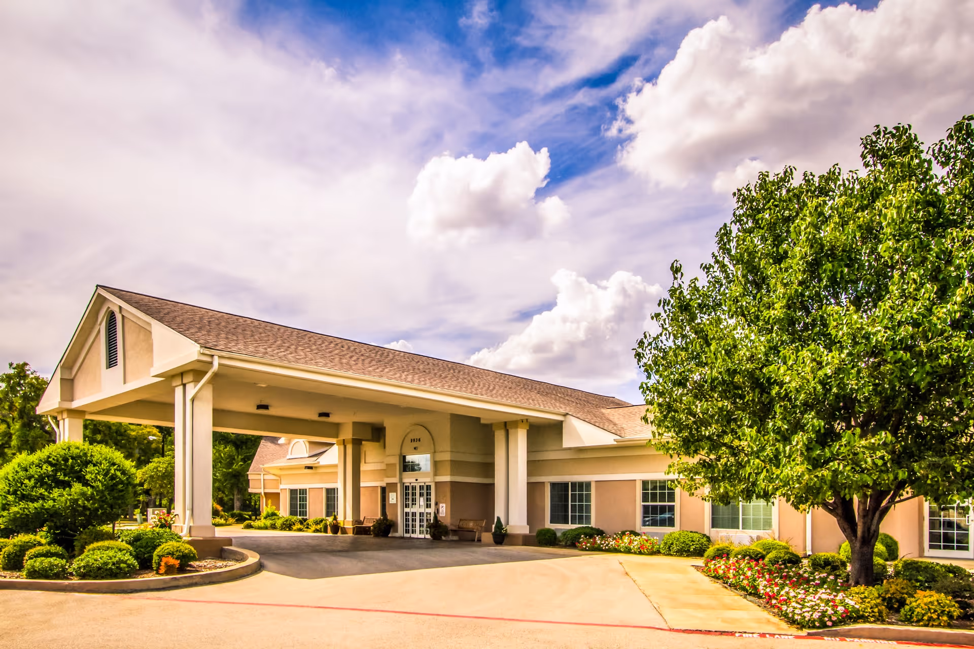 Exterior view of a single-story senior living facility building with a covered entrance, surrounded by well-maintained landscaping including bushes, flowers, and a large tree under a partly cloudy sky.