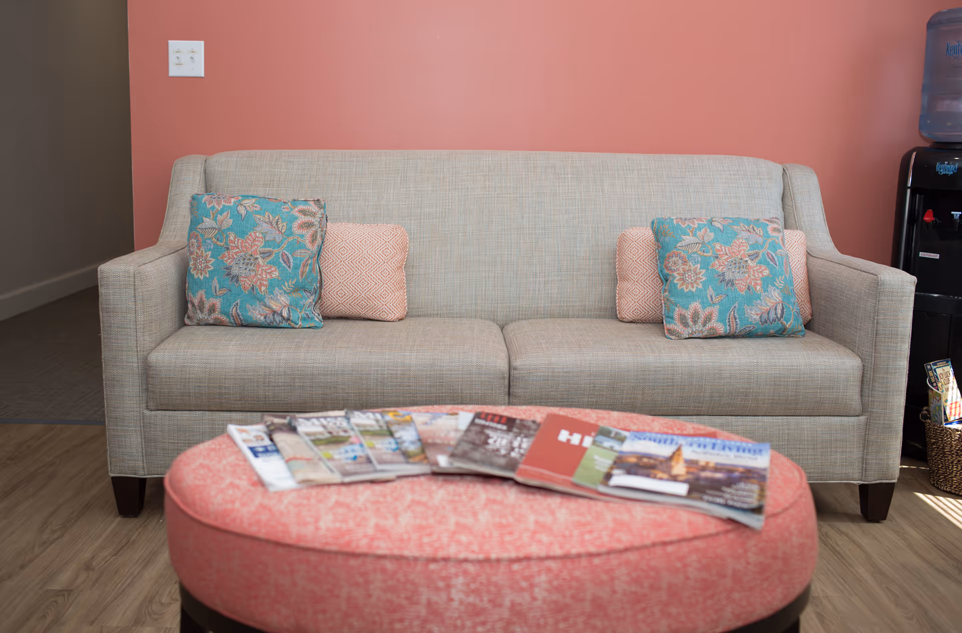 Beige sofa with patterned throw pillows against a coral wall and a pink ottoman with magazines in front.