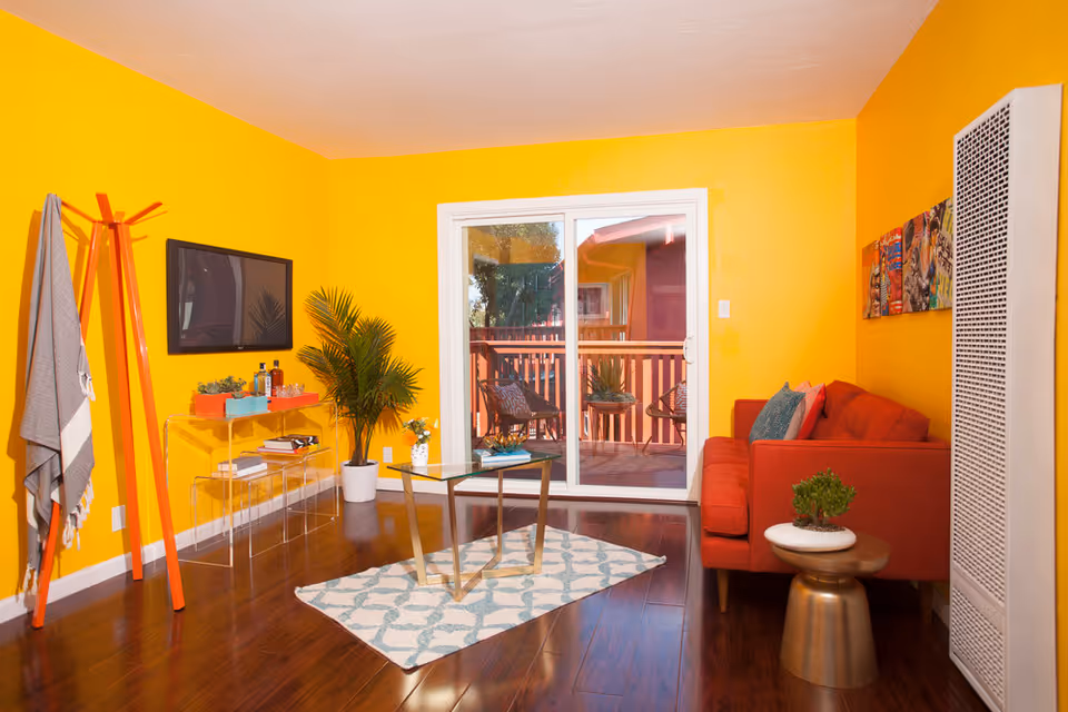A bright living room with yellow walls and dark wooden flooring. The room features a red sofa with colorful cushions, a small round side table with a potted plant, and a glass coffee table on a patterned rug. A flat-screen TV is mounted on the wall above a transparent console table holding books and decorative items. A tall orange coat rack with a gray and white blanket hangs near the TV. A large potted plant is placed next to the console table. Sliding glass doors lead to an outdoor balcony with chairs and a small table. A wall heater is visible on the right side of the room.