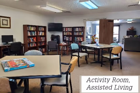 Activity room in an assisted living facility with tables and chairs arranged for group activities. Bookshelves filled with books line the back wall, a television is mounted above a small cabinet, and a computer workstation is visible on the left side. The room is well-lit with fluorescent ceiling lights and has a carpeted floor.