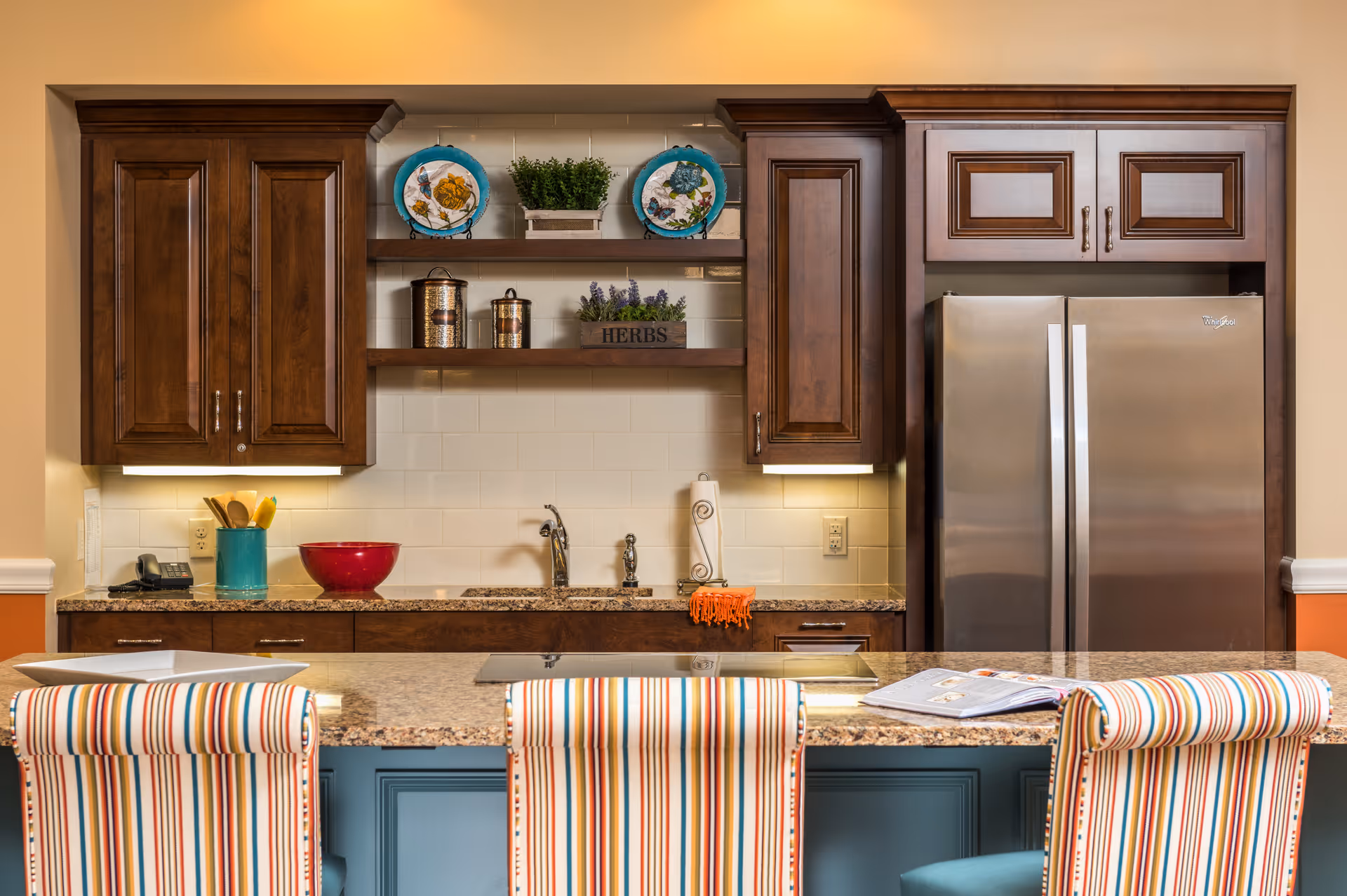 A kitchen area with wooden cabinets, a stainless steel refrigerator, and a granite countertop island with three striped upholstered chairs. The backsplash is white tile, and there are decorative plates, plants, and containers on wooden shelves above the sink.