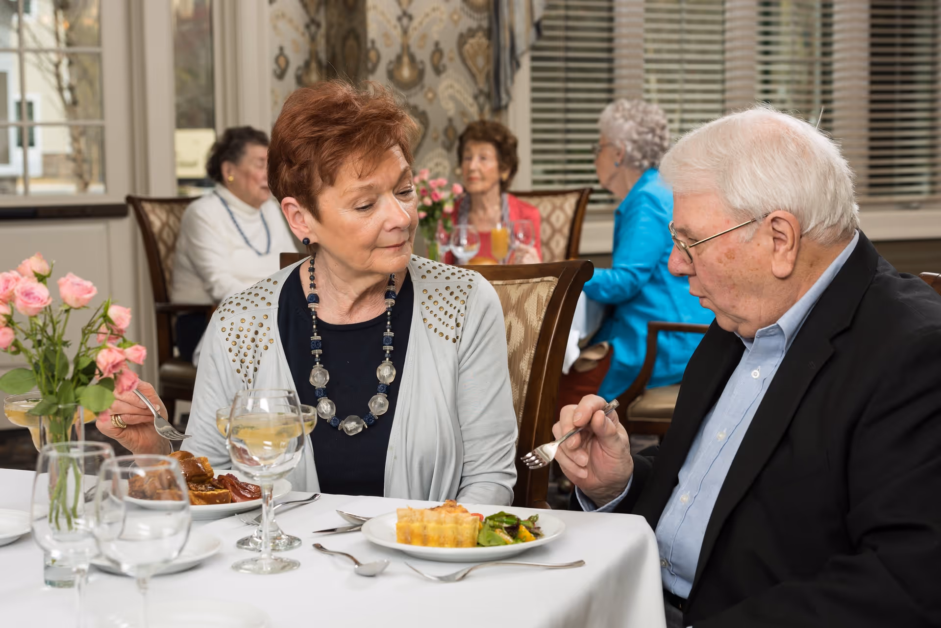 Two elderly individuals seated at a dining table in a well-lit room, enjoying a meal together. The woman on the left is wearing a light gray cardigan and a beaded necklace, while the man on the right is dressed in a dark blazer and light blue shirt. In the background, three other elderly people are seated at another table, engaged in conversation. The table is set with white tablecloths, glassware, and a vase of pink roses.