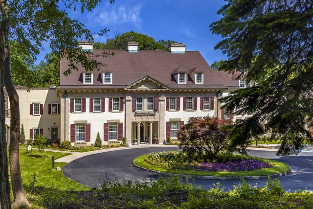 Front exterior view of a large, elegant senior living facility building with cream-colored walls, red shutters, and a brown roof. The building is surrounded by well-maintained landscaping including trees, bushes, and a circular driveway with a flower bed in the center. The sky is clear and blue.