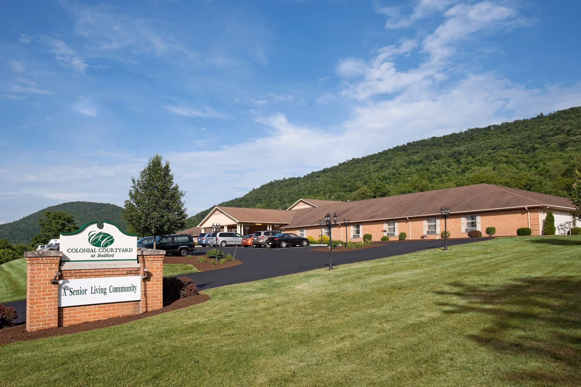 Exterior view of Colonial Courtyard at Bedford, a senior living community, showing a single-story brick building with a brown roof, a parking lot with several cars, a green lawn, and a sign at the entrance. The background features green hills and a partly cloudy blue sky.