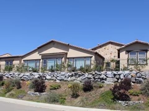 Exterior view of a single-story senior care facility with landscaped hillside and blue sky.