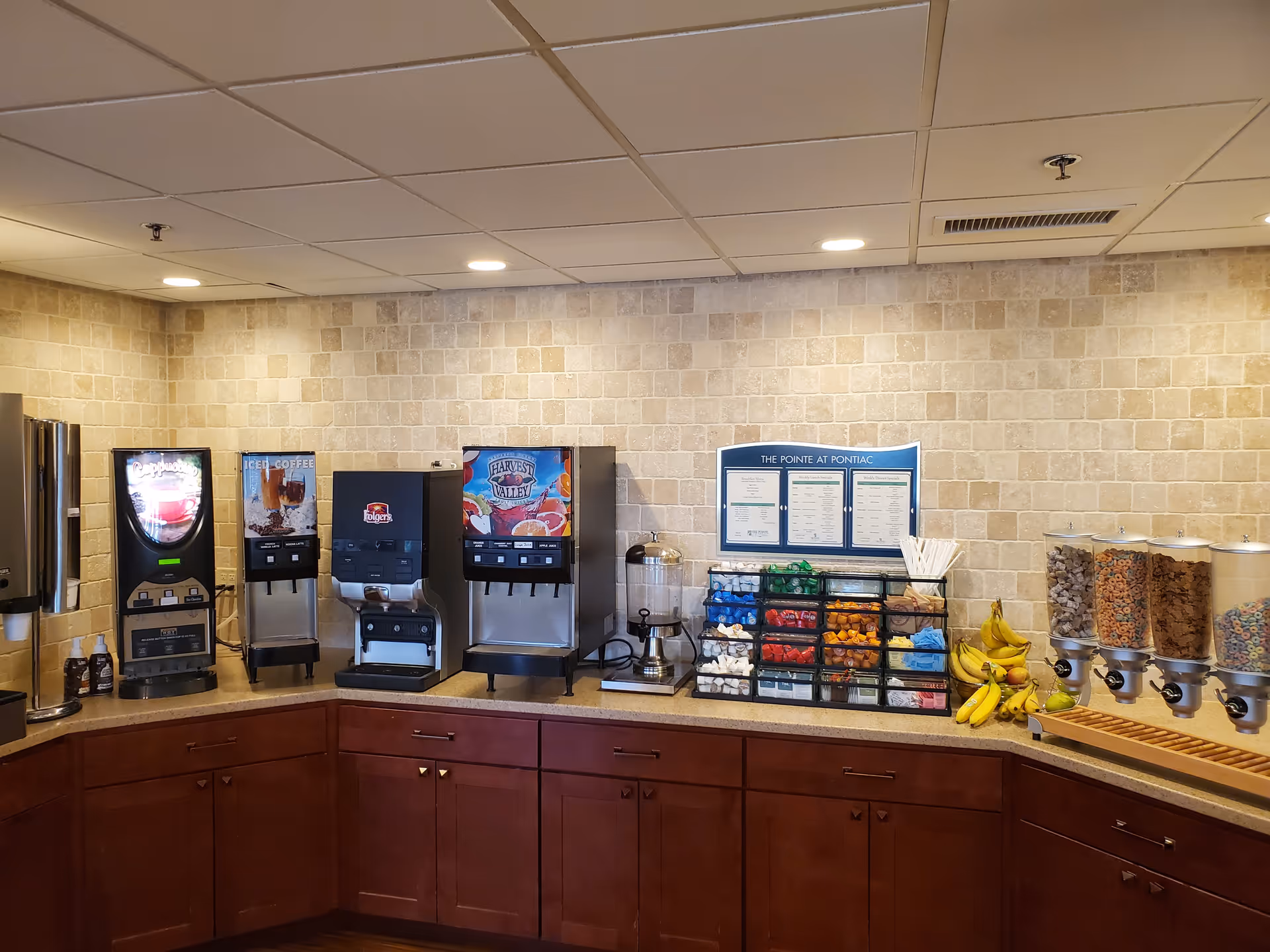 Self-serve breakfast station with coffee machines, cereal dispensers, bananas, and condiment racks along a tiled countertop.