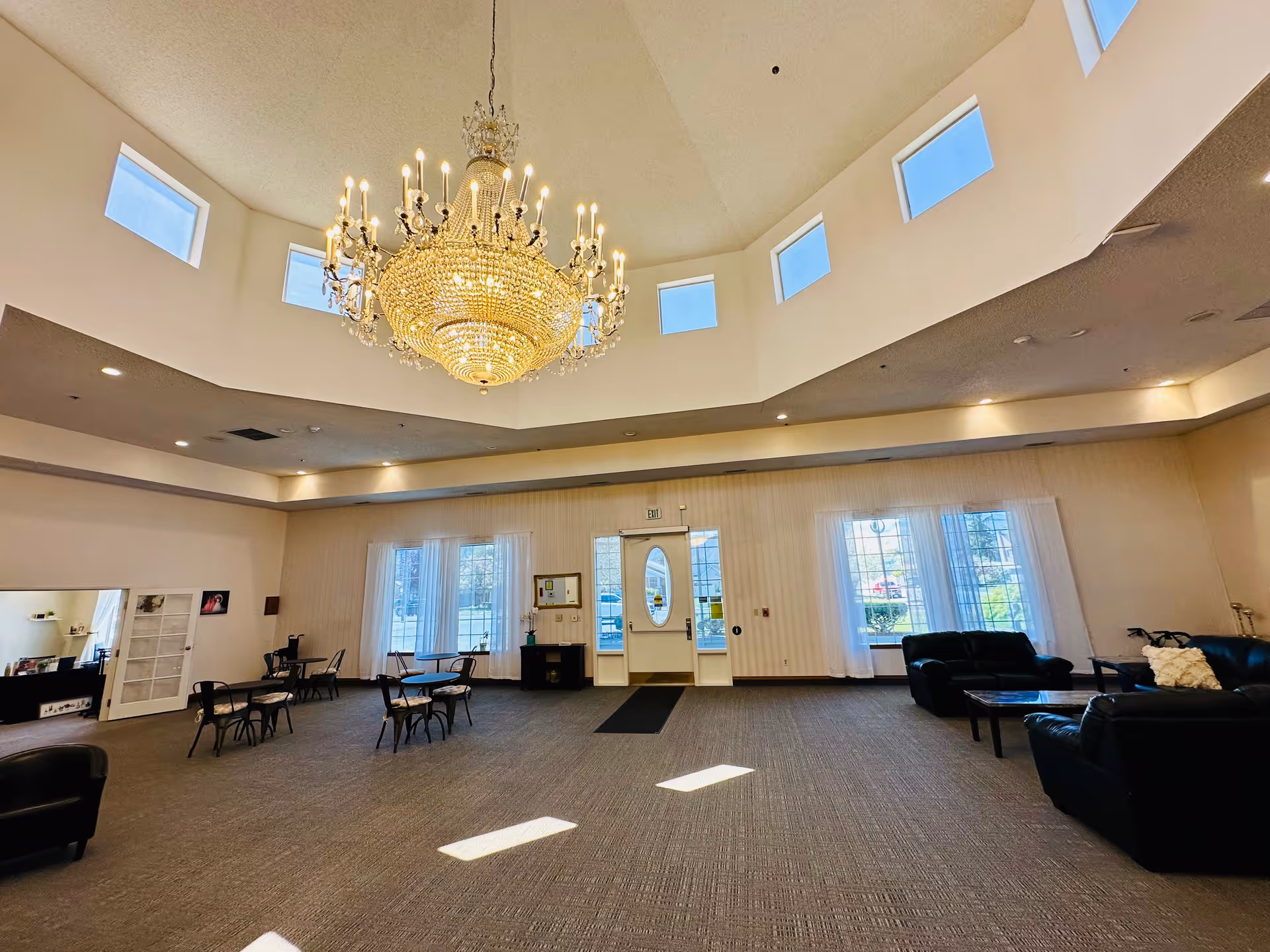 Spacious senior living facility common area with a large ornate chandelier hanging from a high ceiling with multiple small windows. The room features several small tables with chairs, black leather sofas, large windows with sheer white curtains, and a door leading outside. The carpeted floor and soft lighting create a welcoming atmosphere.